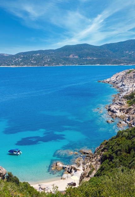Paesaggio marino estivo con barca in acqua trasparente color acquamarina e spiaggia sabbiosa sulla costa rocciosa. Vista dalla riva, Sithonia, Halkidiki, Grecia.