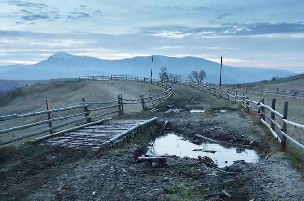 Paesaggio mattutino con strada rurale e pozzanghere. Gelo a terra e recinzione in un villaggio di montagna. Carpazi, Ucraina, Europa