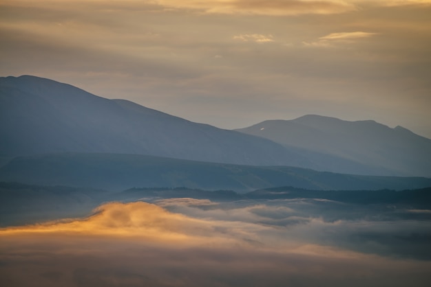 Paesaggio montano panoramico all'alba con nuvole basse dorate nella valle tra sagome di montagne blu sotto il cielo nuvoloso. Tramonto o alba scenario con nuvole basse nella valle di montagna in colori illuminanti.