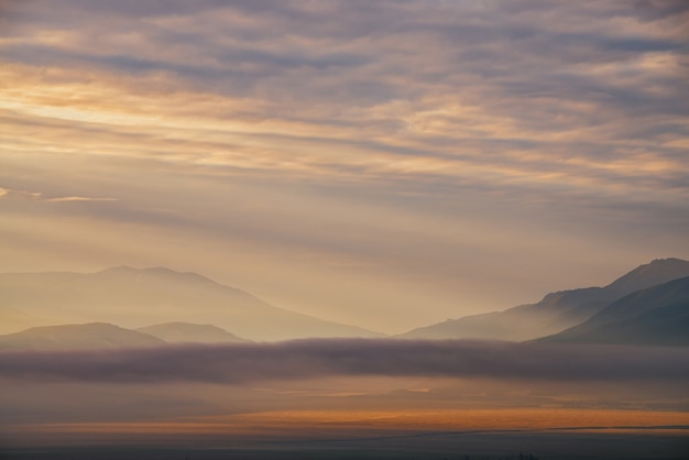 Paesaggio montano panoramico all'alba con nuvole basse dorate nella valle tra sagome di montagne sotto il cielo nuvoloso. Vivido scenario al tramonto o all'alba con nuvole basse nella valle di montagna in colori illuminanti.