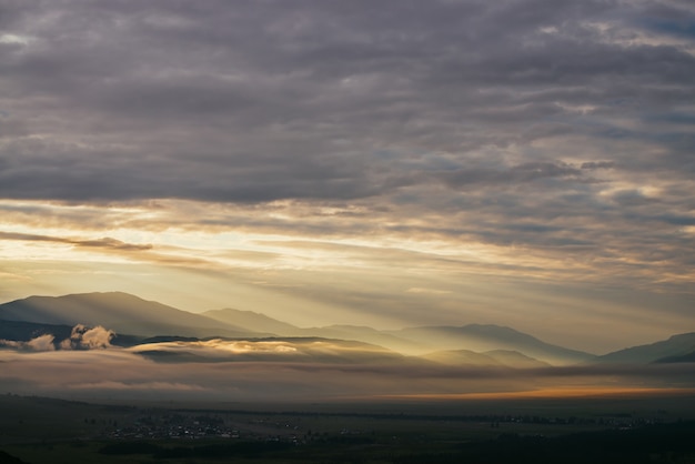 Paesaggio montano panoramico con nuvole basse dorate sopra il villaggio tra sagome di montagne sotto il cielo nuvoloso dell'alba. Atmosferico scenario alpino di campagna in nuvole basse al tramonto illuminante a colori.