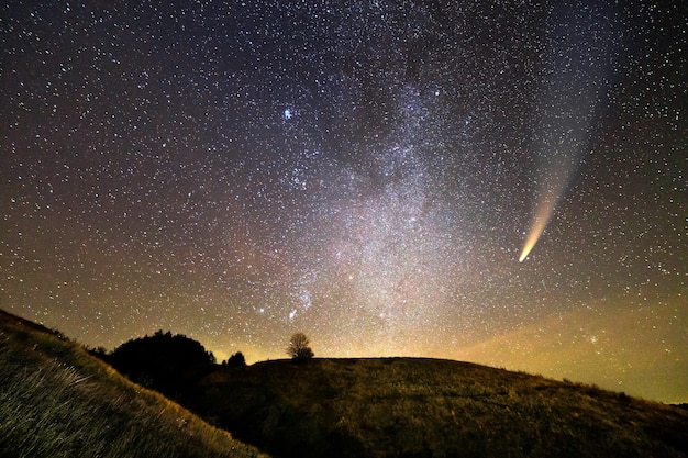 Paesaggio notturno di montagne con cielo coperto di stelle e cometa Neowise con coda leggera.