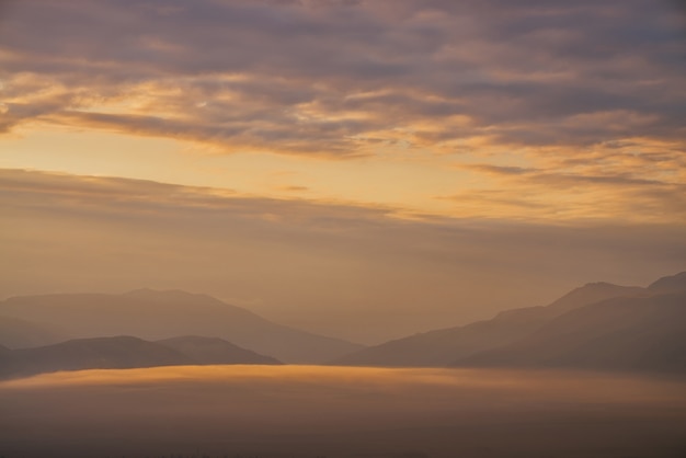 Paesaggio panoramico all'alba con nuvole basse dorate nella valle tra sagome di montagne sotto il cielo nuvoloso.