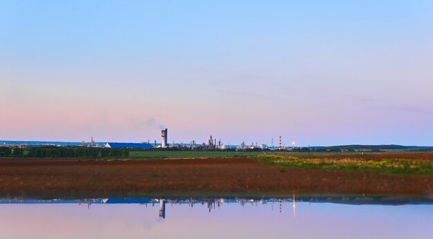 Paesaggio rurale con un impianto chimico all'orizzonte riflesso nell'acqua in primo piano sfocato