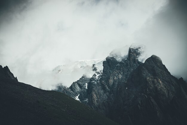 Paesaggio surreale atmosferico scuro con cima di montagna rocciosa scura in nuvole basse in cielo nuvoloso grigio. Nuvola bassa grigia sull'alto pinnacolo. Alta roccia nera con neve in nuvole basse. Montagne cupe surrealiste.