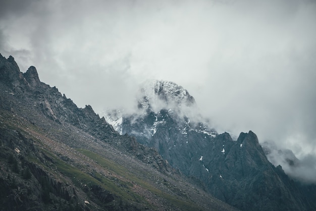 Paesaggio surreale atmosferico scuro con cima di montagna rocciosa scura in nuvole basse in cielo nuvoloso grigio. Nuvola grigia bassa sull'alto pinnacolo. Alta roccia nera con neve in nuvole basse. Montagne cupe surrealiste.