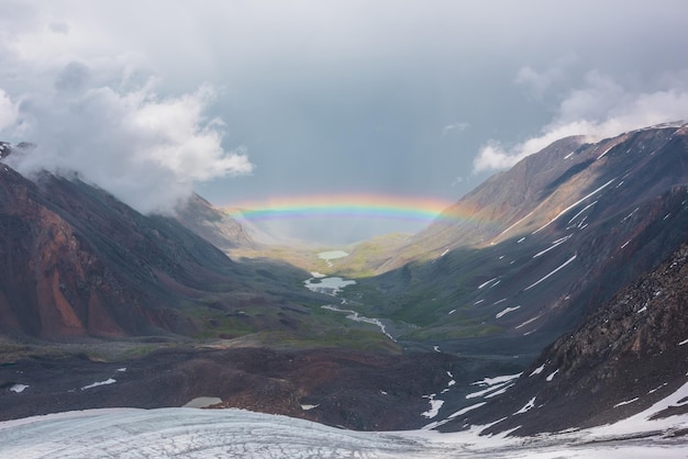 Paesaggio tetro con un vivido arcobaleno sopra il lago di montagna Paesaggio cupo con un arcobaleno luminoso sopra il ghiacciaio e il lago glaciale nella valle di montagna Vista dall'alto dell'arcobaleno colorato e delle nuvole basse in montagna