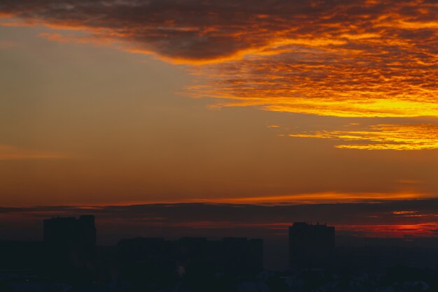 Paesaggio urbano con vivida alba infuocata. Cielo nuvoloso drammatico caldo stupefacente sopra le siluette scure dei tetti della costruzione della città. Luce solare arancione.