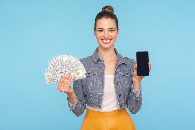 Pagamento mobile, banking online. Ritratto di donna vestita alla moda allegra carina con panino per capelli che tiene cellulare e banconote da un dollaro, sorridendo con gioia. tiro in studio indoor isolato su sfondo blu