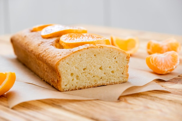 Pagnotta di deliziosa torta al mandarino su pergamena con pezzi di mandarino su fondo rustico in legno Fetta di torta agli agrumi secondo ricetta classica Dessert vegano da forno fatto in casa