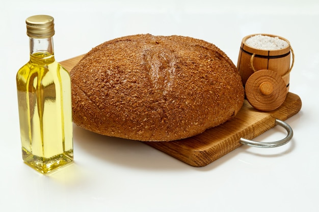 Pagnotta di pane di segale su tagliere di legno, una bottiglia di olio di girasole e una cantina di sale in legno con la superficie bianca