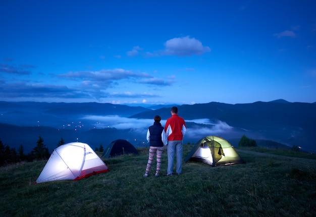 Paia attive di retrovisione che si tengono per mano godendo dell'alba vicino al campeggio nelle montagne. Colline nebbiose e cielo blu creano uno splendido paesaggio mattutino