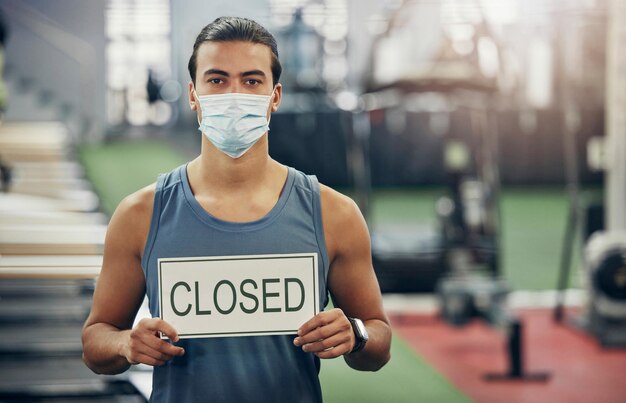 Palestra Covid e uomo con cartello chiuso durante la pandemia mentre indossa una maschera Centro sanitario per coronavirus e blocco della quarantena con maschio forte in forma in palestra vuota