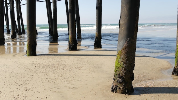 Pali di legno sotto il lungomare, il vecchio molo di Oceanside, costa della California USA. Piloni, piloni o pilastri sotto il ponte vintage retrò, passeggiata sul lungomare. Onde dell'oceano, marea dell'acqua di mare e spiaggia di sabbia.