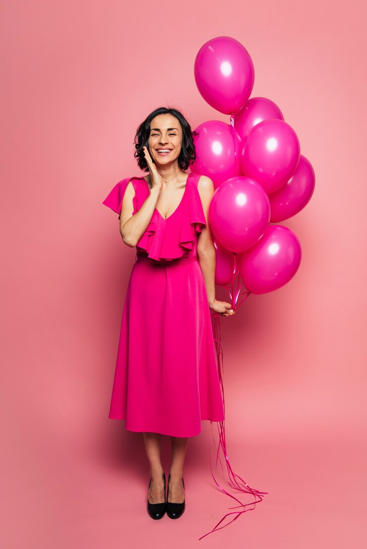 Foto palloncini rosa. foto a figura intera di una ragazza felice in un lungo abito fucsia, che sorride con gli occhi chiusi e tiene dei palloncini rosa nella mano sinistra.