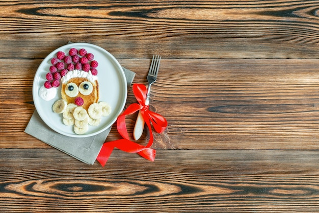 Pancake a forma di Babbo Natale di Natale con bacche di lampone fresco dolce e banana sul piatto su fondo di legno per la colazione dei bambini dei bambini. cibo di natale con decorazioni di capodanno con spazio di copia.