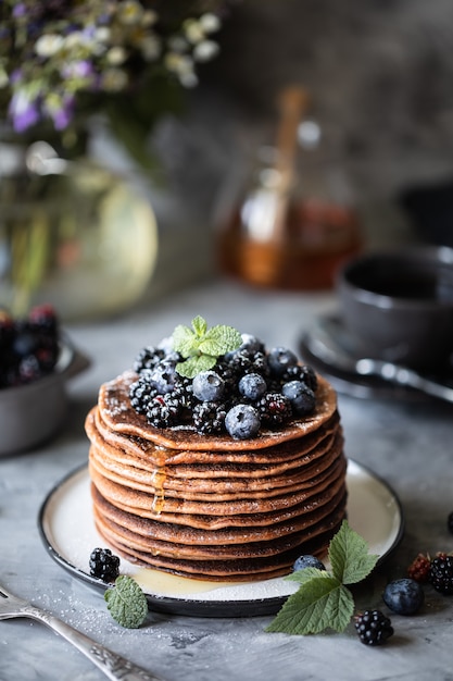 Pancake al cioccolato con frutti di bosco e frutta con miele, con zucchero a velo volante e un mazzo di fiori selvatici sul tavolo. Buio