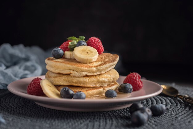 Pancake con fragole, lamponi, mirtilli, banana e sciroppo su un piatto rosa.