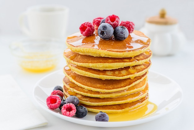 Pancake con miele, mirtilli e lamponi su un piatto bianco.