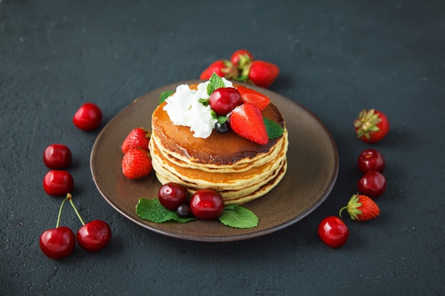 Pancake in un piatto con fragole, panna montata, menta, miele e ciliegia su uno sfondo nero scuro