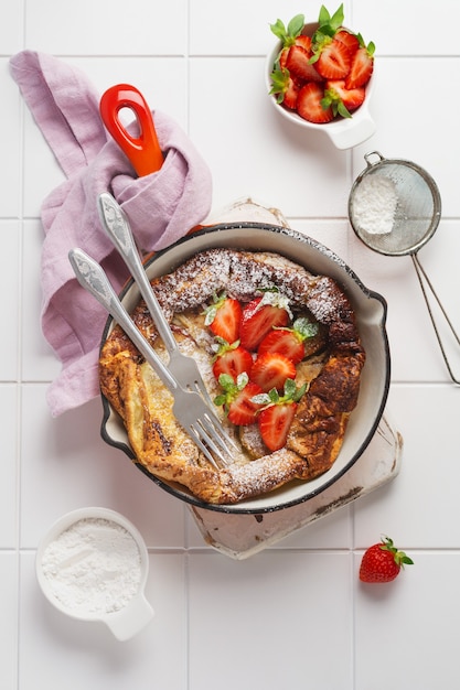 Pancake olandese del bambino con la bacca fresca della fragola e cosparso di polvere di zucchero a velo in padella rossa sul muro bianco della cucina. Vista dall'alto.