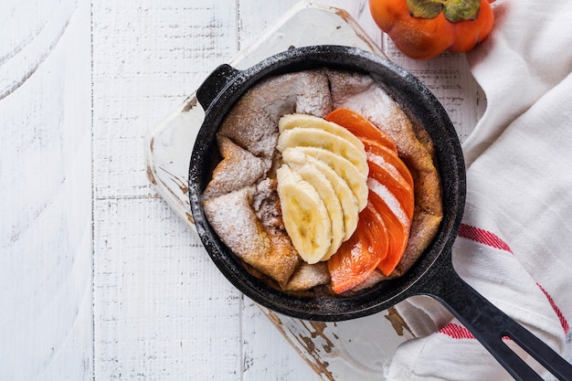 Pancake olandese del bambino con la mela, il cachi, la banana, la cannella in una piccola padella del ferro sulla superficie del tavolo in legno