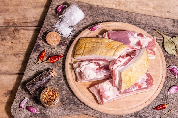 Pancetta di suino cruda con cotenna, carne di peritoneo. Spezie ed erbe aromatiche, ingredienti per cucinare pasti sani, proteine animali. Tavolo vintage in legno, vista dall'alto