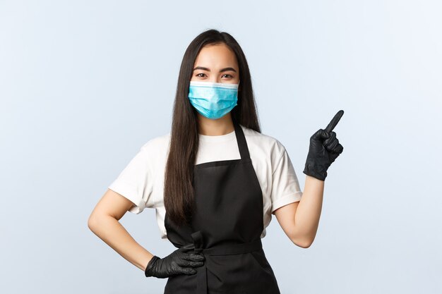 Pandemia di Covid-19, distanza sociale, piccole imprese e prevenzione del concetto di virus. Sorridente barista asiatico della caffetteria, impiegato in maschera medica e guanti che puntano l'angolo in alto a destra.