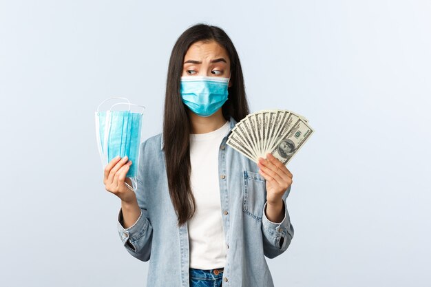 Pandemia di Covid-19, spese di coronavirus e concetto di finanza. Donna asiatica premurosa e perplessa che guarda la maschera medica e mostra soldi, perdendo soldi durante il coronavirus