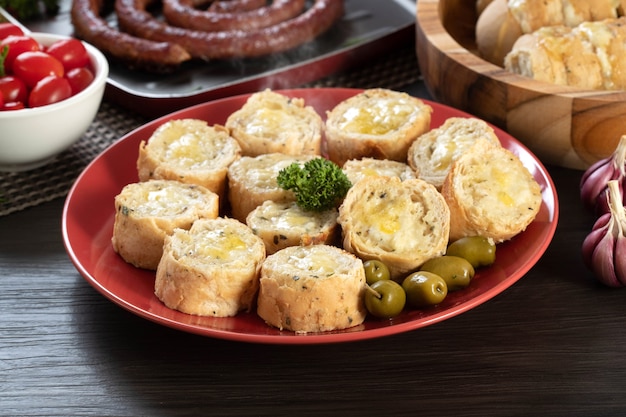 Pane all'aglio su un piatto rosso sul tavolo del barbecue con salsiccia, formaggio, rosmarino, olive e pomodorini.