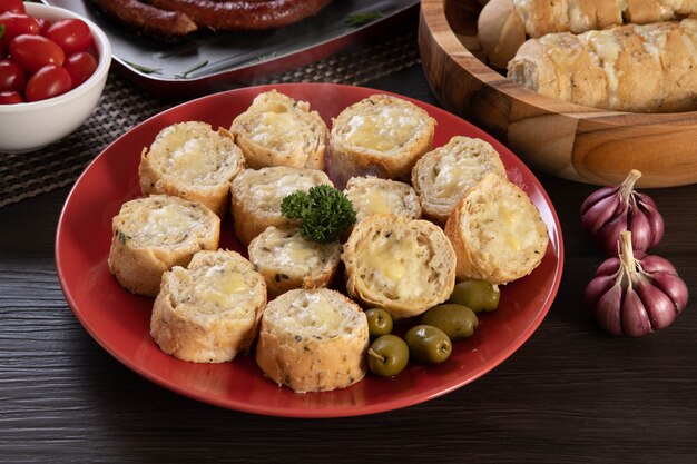 Pane all'aglio su un piatto rosso sul tavolo del barbecue con salsiccia, formaggio, rosmarino, olive e pomodorini.