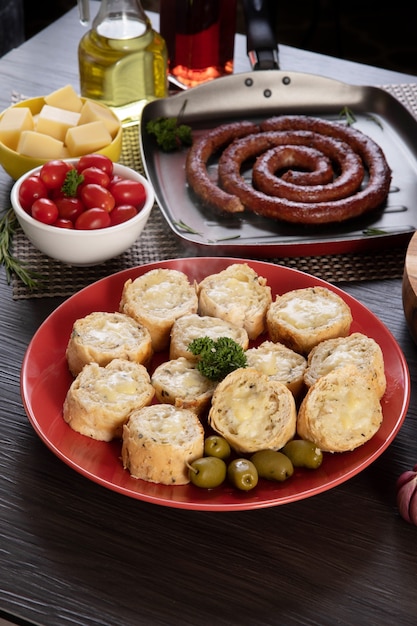 Pane all'aglio su un piatto rosso sul tavolo del barbecue con salsiccia, formaggio, rosmarino, olive e pomodorini.