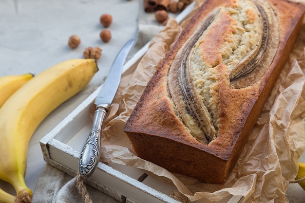 Pane alla banana fatto in casa fresco in vassoio di legno bianco con ingredienti su cemento leggero.