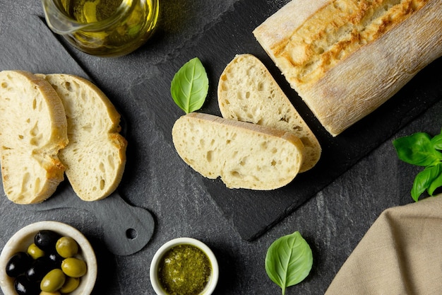 Pane ciabatta italiano fresco con erbe aromatiche, olio d'oliva, olive nere e verdi, foglie di basilico e salsa al pesto su sfondo scuro. Vista dall'alto.