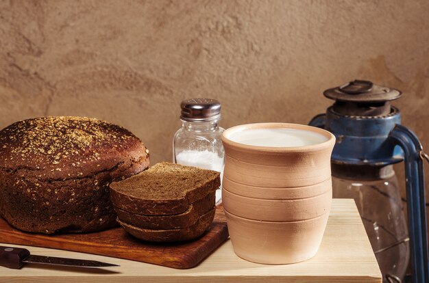 Pane di segale con sale su un tagliere, pentola di ceramica con latte e una lanterna a cherosene. Natura morta in stile rustico