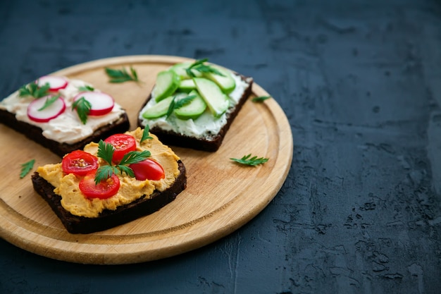 Pane di segale vegetariano sano panini aperti con ricotta, hummus, avocado, ravanello e pomodoro. Tavola di legno su una superficie nera, copia-spazio