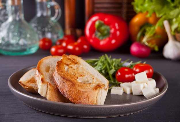 Pane tostato a fette sulla piastra nera con pomodori, feta e rucola con verdure sullo sfondo