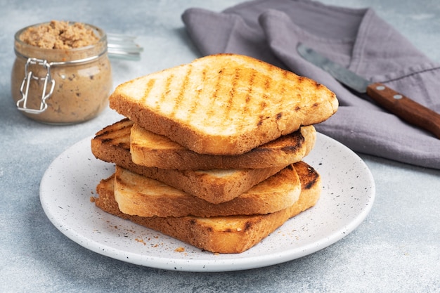 Pane tostato croccante per panini su un piatto su un tavolo di cemento grigio. Un vasetto di patè di pollo.