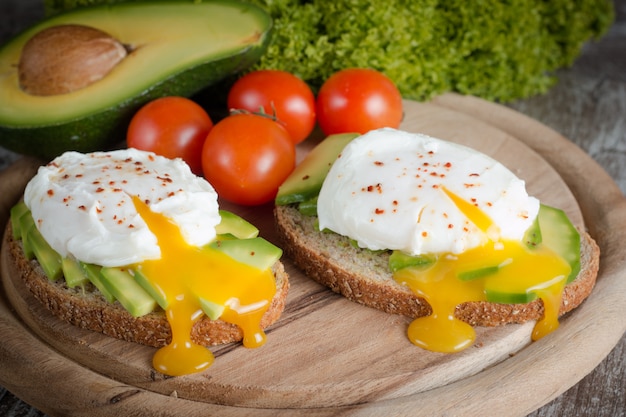 Pane tostato dell&#39;avocado, pomodoro ciliegia e uova affogate su fondo di legno.