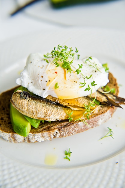 Pane tostato di pane nero con avocado, uovo in camicia e spratti.