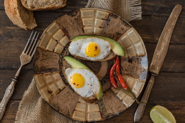 Pane tostato rurale della prima colazione con le uova fritte e l'avocado sul bordo di legno sopra fondo marrone, vista superiore.