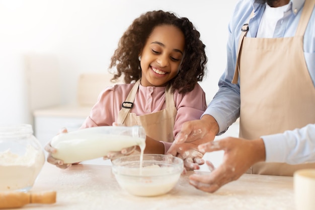 Panetteria casalinga. Primo piano della sorridente ragazza afroamericana che versa il latte in una ciotola di vetro con pasta per biscotti, cucina insieme a papà, prepara una torta per celebrare la festa del papà, il fuoco selettivo