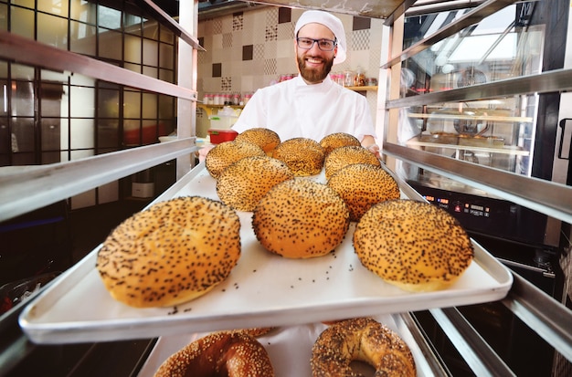 Panettiere bello in uniforme bianca che tiene un vassoio con le pasticcerie fresche di una fabbrica del pane o del forno