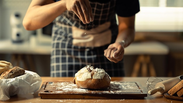 Panettiere maschio che indossa il grembiule setacciando la farina sull'impasto che fa il pane in cucina