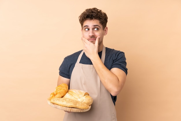 Panettiere maschio che tiene una tavola con parecchi pani su beige che pensa un'idea