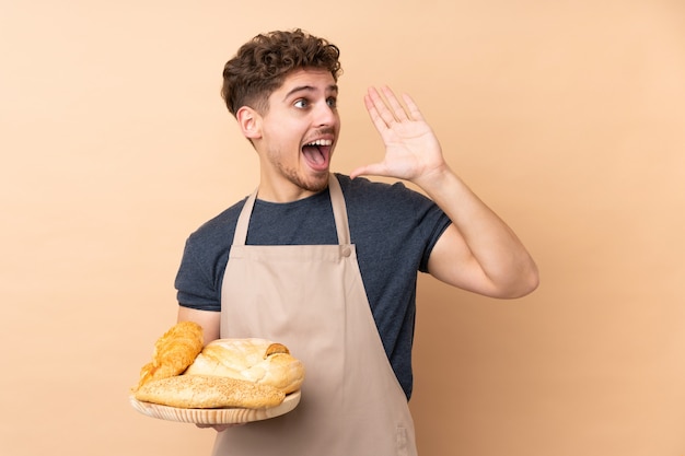 Panettiere maschio che tiene una tavola con parecchi pani sulla parete beige che grida con la bocca spalancata