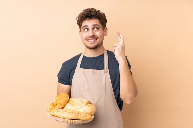 Panettiere maschio che tiene una tavola con parecchi pani sulla parete beige con l'incrocio delle dita