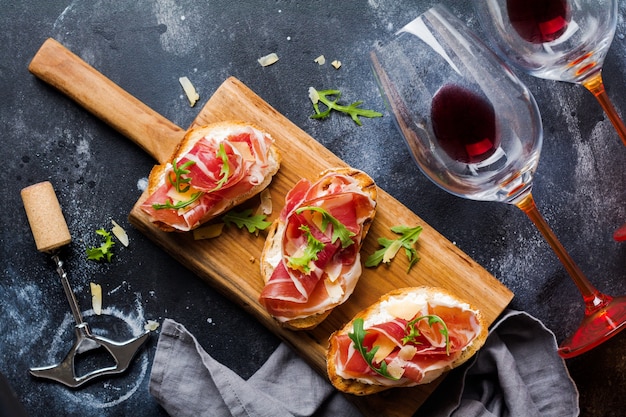 Panini aperti al prosciutto, rucola e formaggio a pasta dura, serviti su supporto in legno con un bicchiere di vino rosso su una vecchia superficie scura di cemento