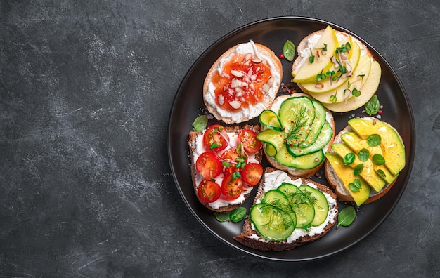 Panini assortiti con formaggio a pasta molle, verdure e frutta su fondo scuro