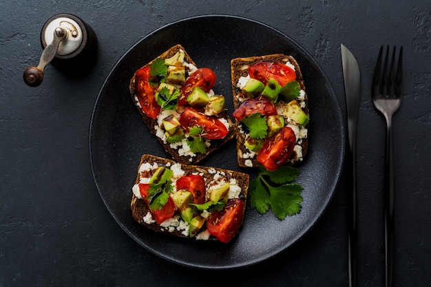 Panini con avocado, pomodorini e feta con salsa balsamica e coriandolo su superficie nera scura. Vista dall'alto.